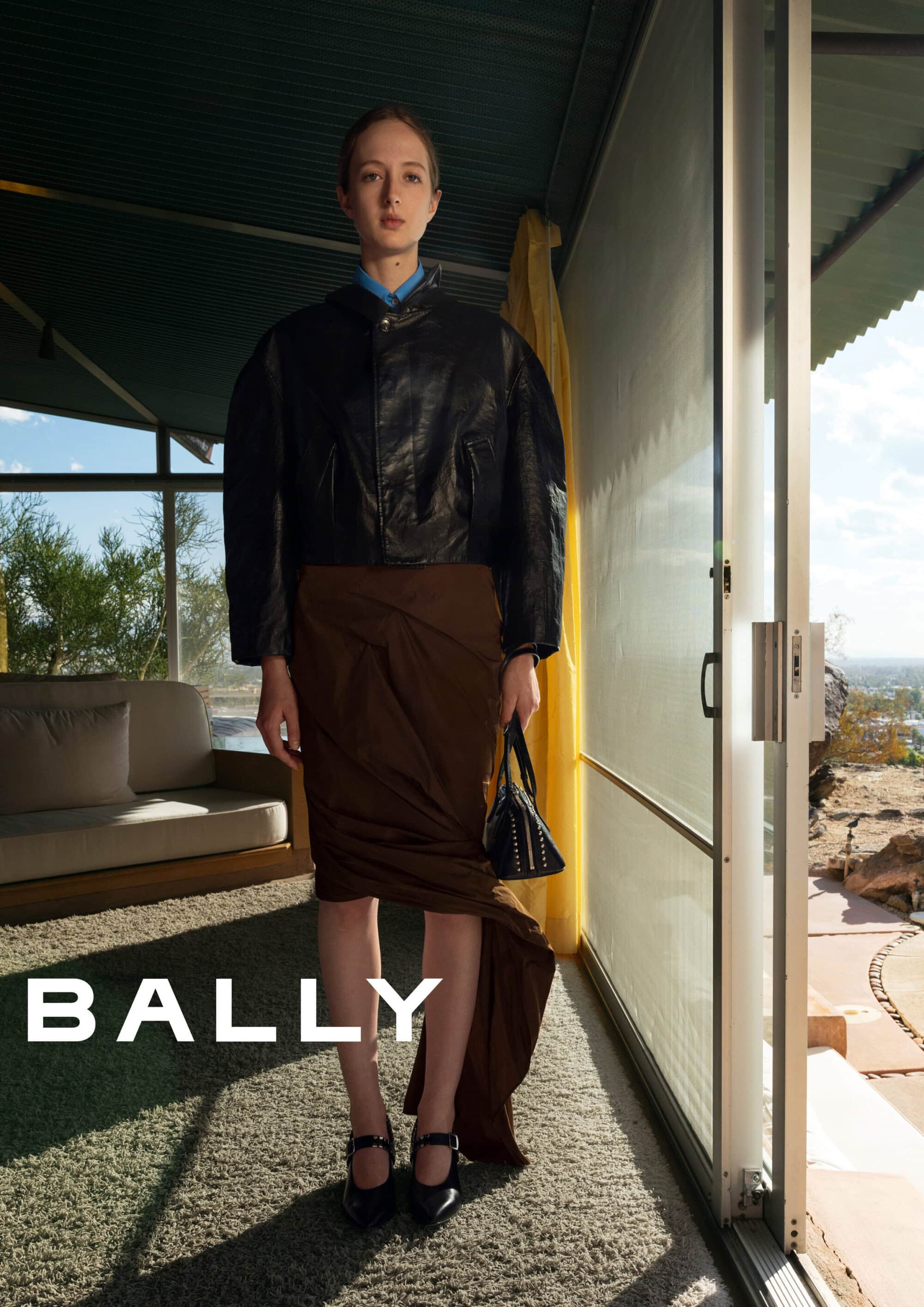 Model standing indoors wearing a black leather jacket and brown draped skirt holding a black handbag for Bally.
