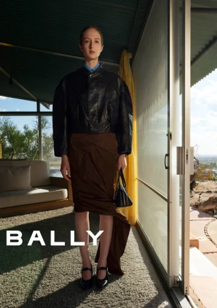 Model standing indoors wearing a black leather jacket and brown draped skirt holding a black handbag for Bally.