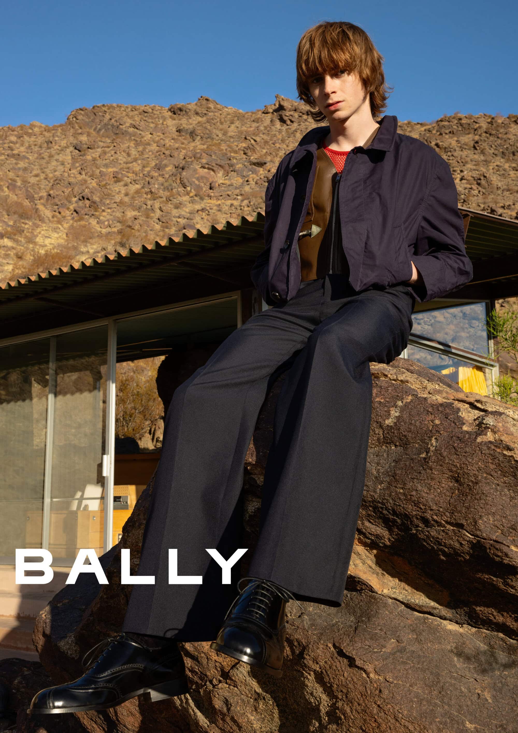 Young male model seated on large rock wearing dark jacket and trousers with desert and modern building in background