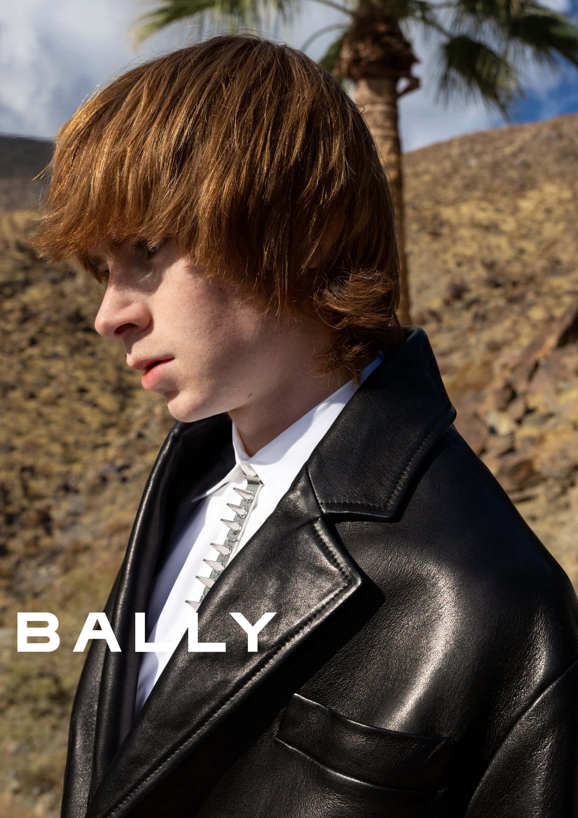 Close-up portrait of a male model wearing a black leather jacket and white shirt outdoors against a desert hillside and palm tree.