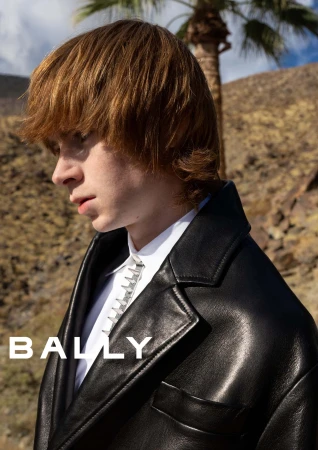 Close-up portrait of a male model wearing a black leather jacket and white shirt outdoors against a desert hillside and palm tree.