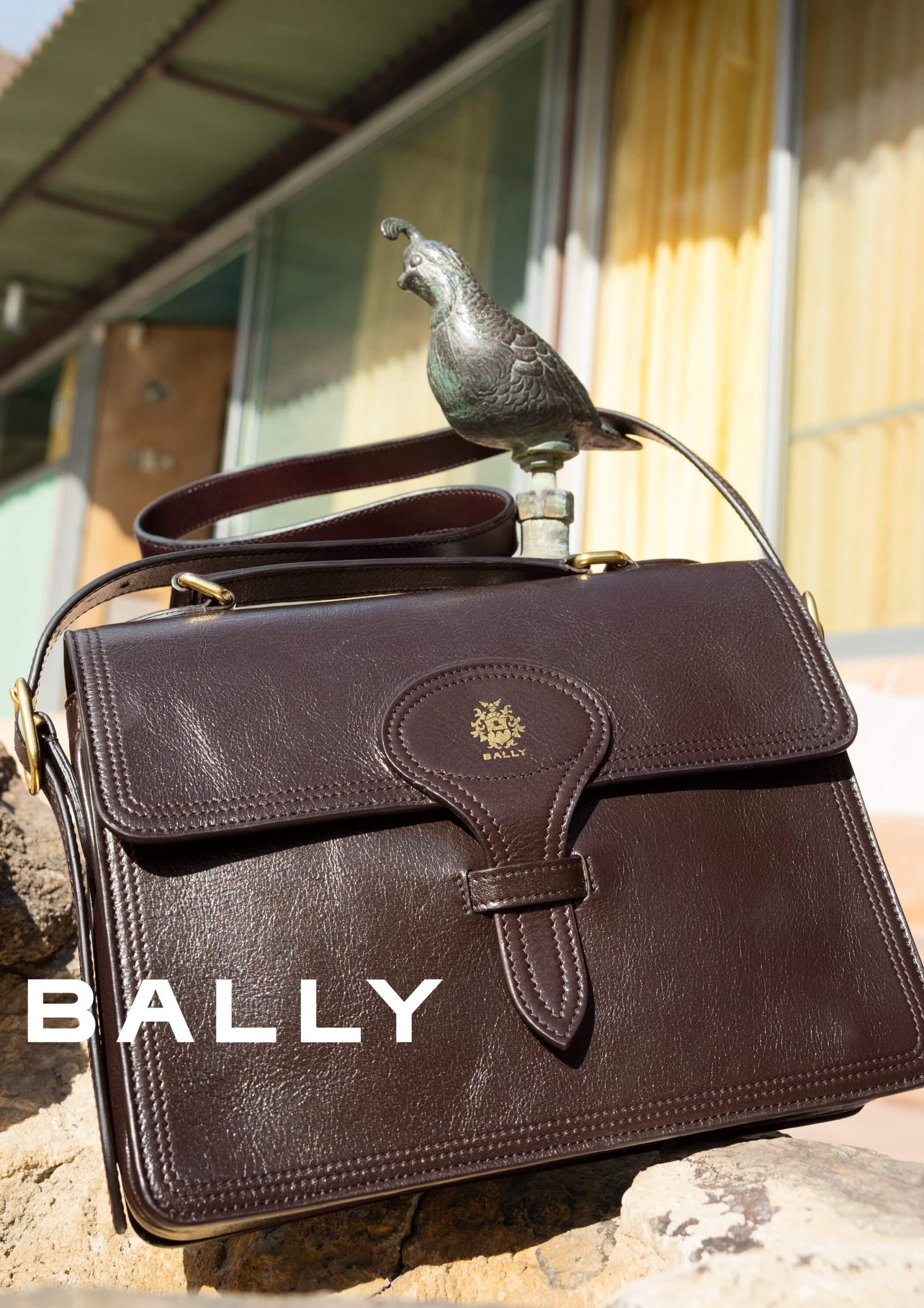 Dark brown leather shoulder bag resting on a stone surface with a small bronze bird figurine perched above it