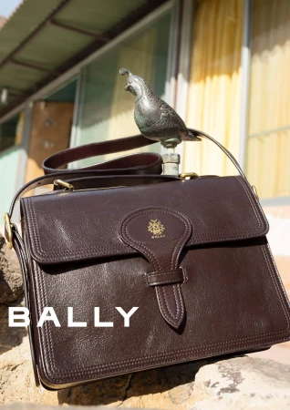 Dark brown leather shoulder bag resting on a stone surface with a small bronze bird figurine perched above it