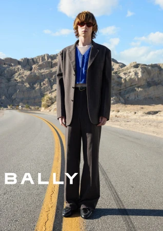 Model standing on a desert road wearing a brown suit jacket, blue zip top, and sunglasses for Bally Spring 2025 campaign