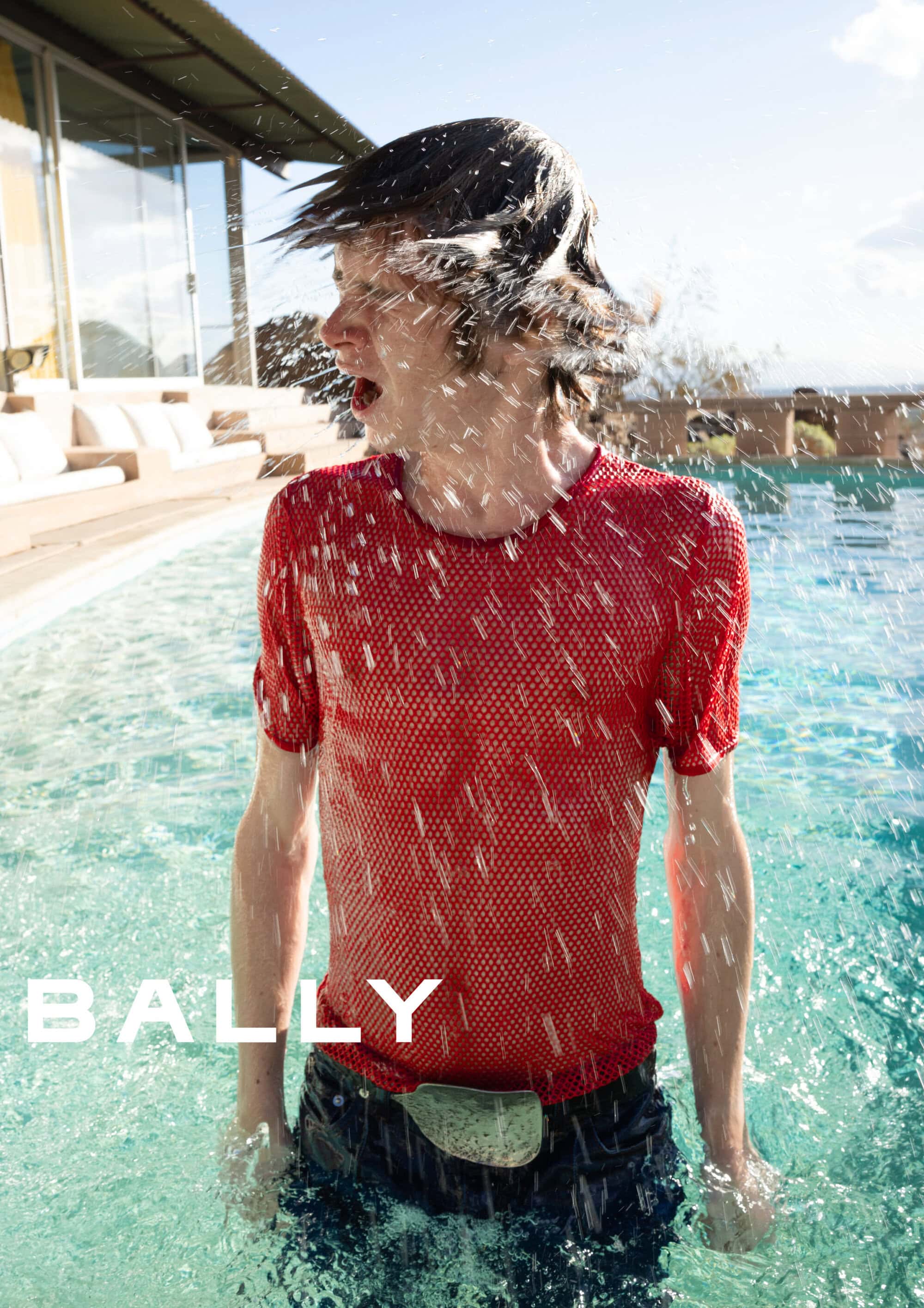 A man wearing a red mesh shirt standing in a swimming pool while water splashes across his face.