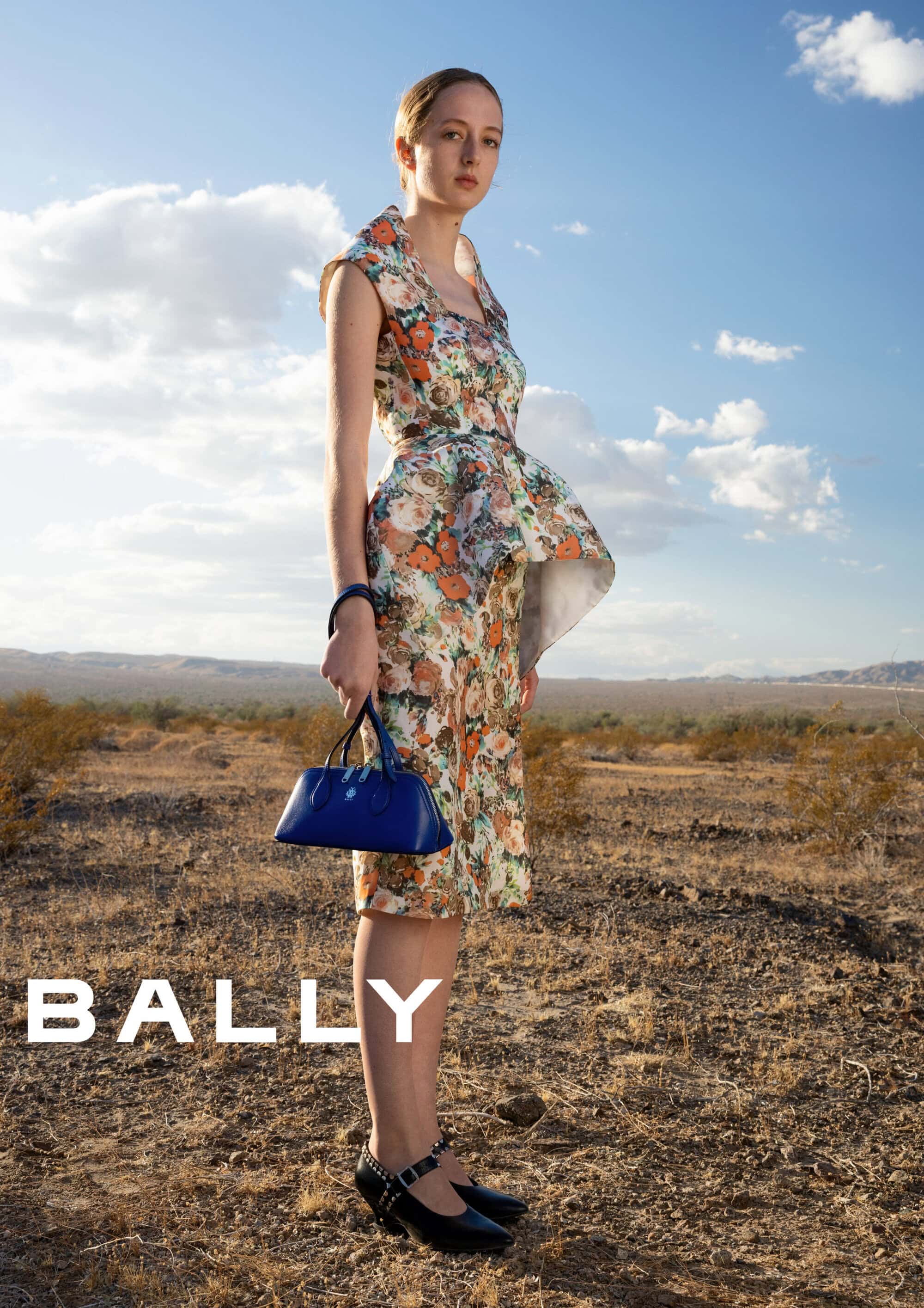Woman wearing a floral patterned dress holding a blue handbag outdoors in a desert landscape under a cloudy blue sky.