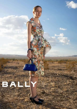 Woman wearing a floral patterned dress holding a blue handbag outdoors in a desert landscape under a cloudy blue sky.