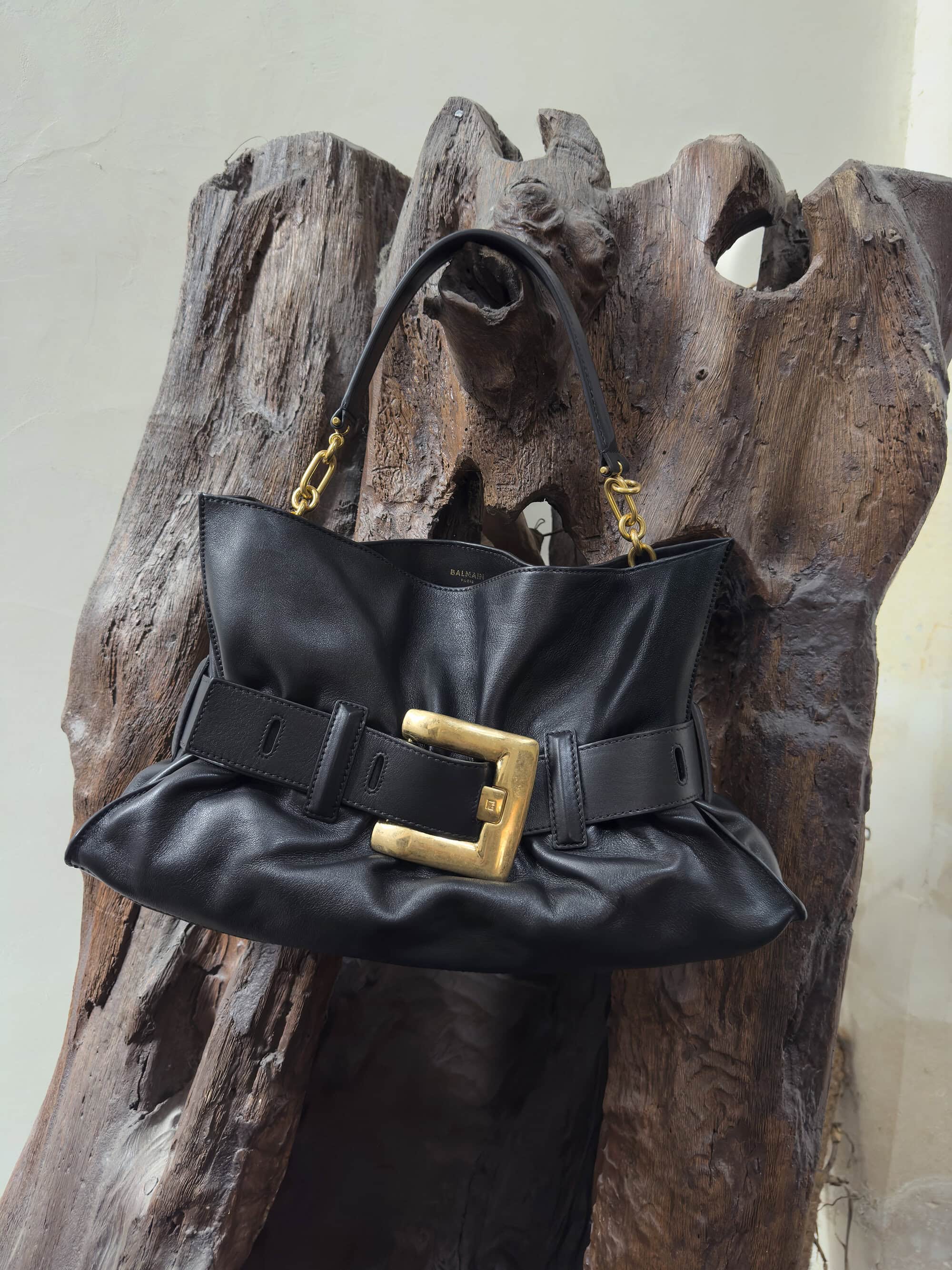 A black leather handbag with a large gold buckle hangs on a piece of rustic dark wood.