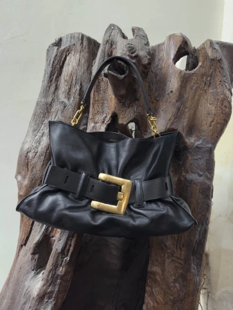 A black leather handbag with a large gold buckle hangs on a piece of rustic dark wood.