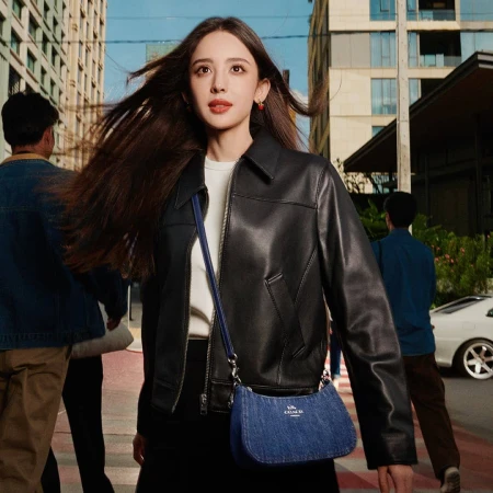 Woman with long hair wearing a black leather jacket and carrying a blue denim crossbody bag on a city street.