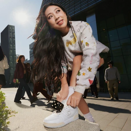 Youngji Lee tying white sneakers with dice accessory wearing patterned sweatshirt outdoors for Coach Spring 2025 campaign