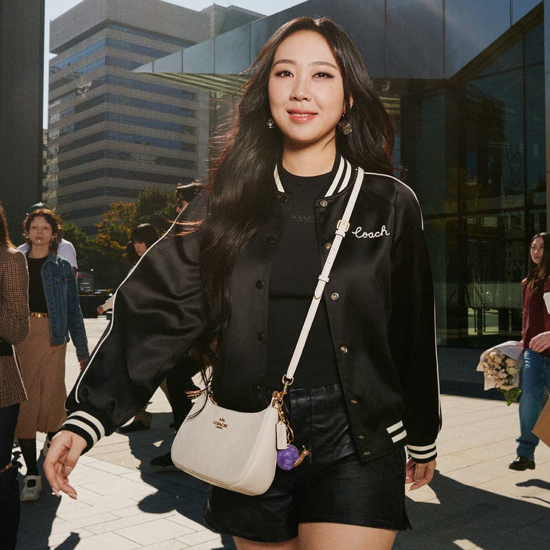 Youngji Lee wearing a black varsity jacket and white crossbody bag outdoors in an urban square