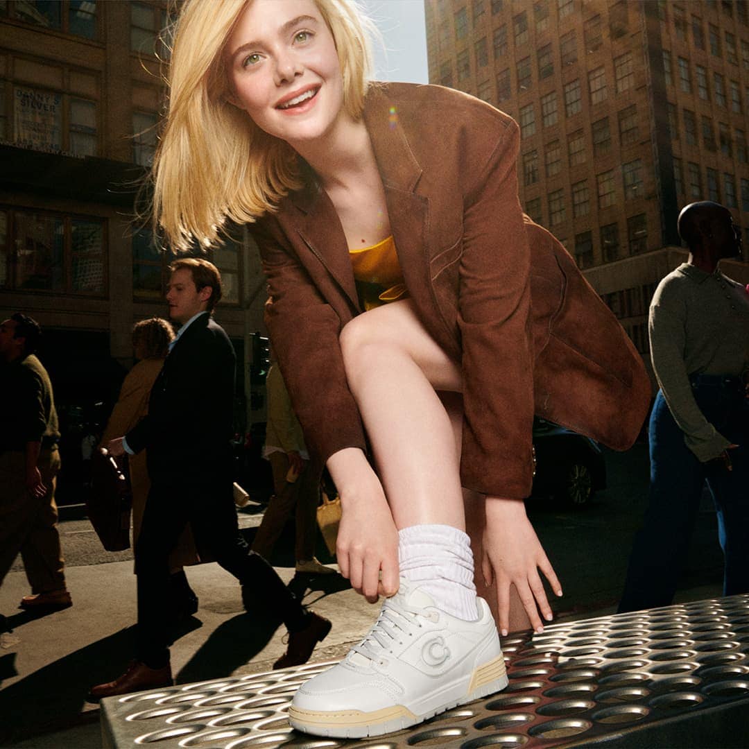 Elle Fanning adjusting her white sneakers outdoors against an urban street background for the Coach Spring 2025 campaign.