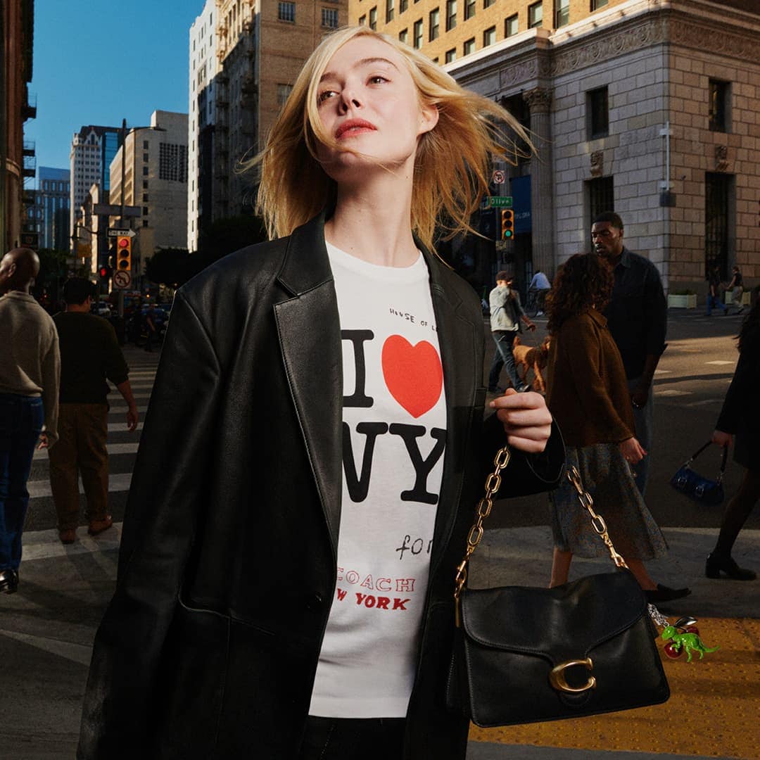 Elle Fanning wearing a black leather blazer and I Love NY t-shirt carrying a black shoulder bag on a city street.