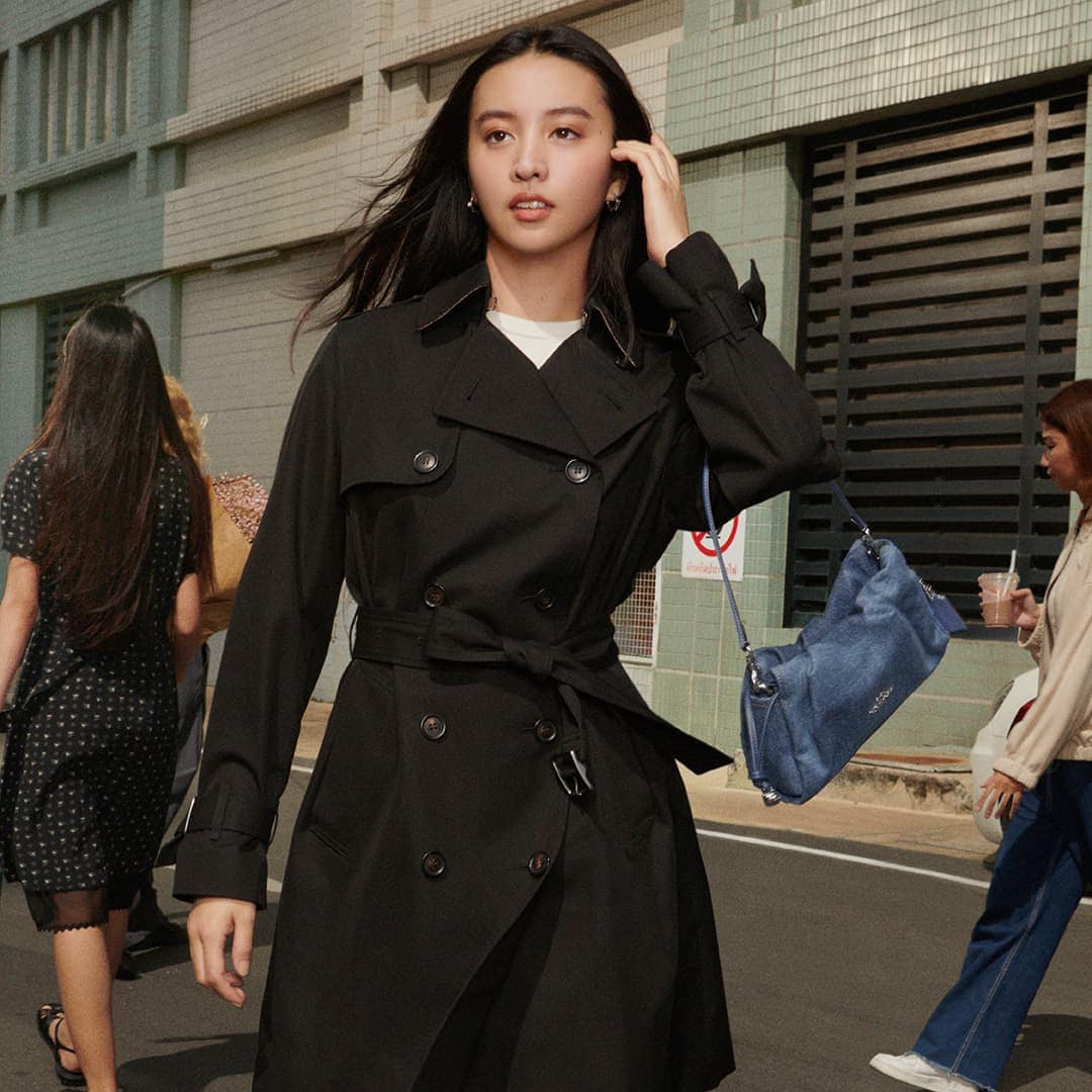 Kōki walking outdoors wearing a black trench coat and carrying a blue handbag for the Coach Spring 2025 ad campaign.
