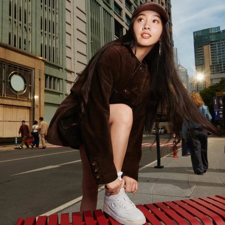 Young woman with long dark hair tying her white sneaker on a city street, wearing a brown jacket and matching cap.