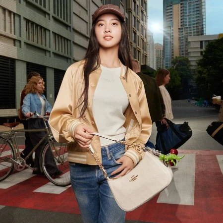 Young woman in a satin bomber jacket and cap holding a white shoulder bag on a city street