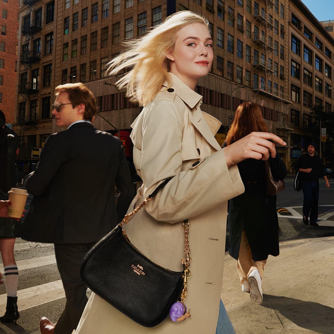 Elle Fanning in a beige trench coat walking on an urban street holding a black shoulder bag for Coach Spring 2025 campaign.