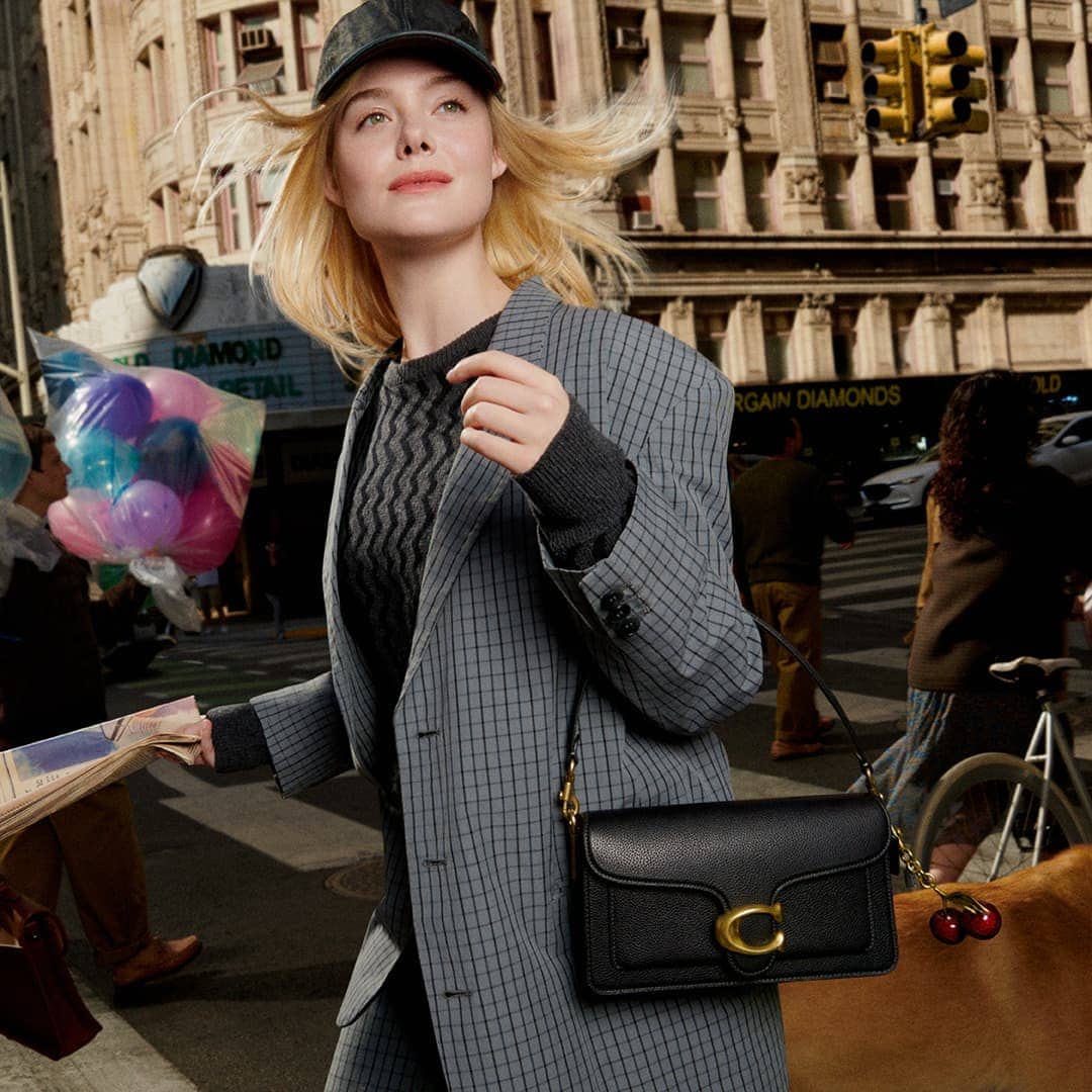 Elle Fanning wearing a grey plaid blazer and baseball cap carrying a black leather shoulder bag in a city street