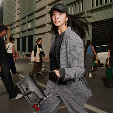 Kōki wearing a grey plaid suit and black cap holding a black handbag with cherry charms on a city street.