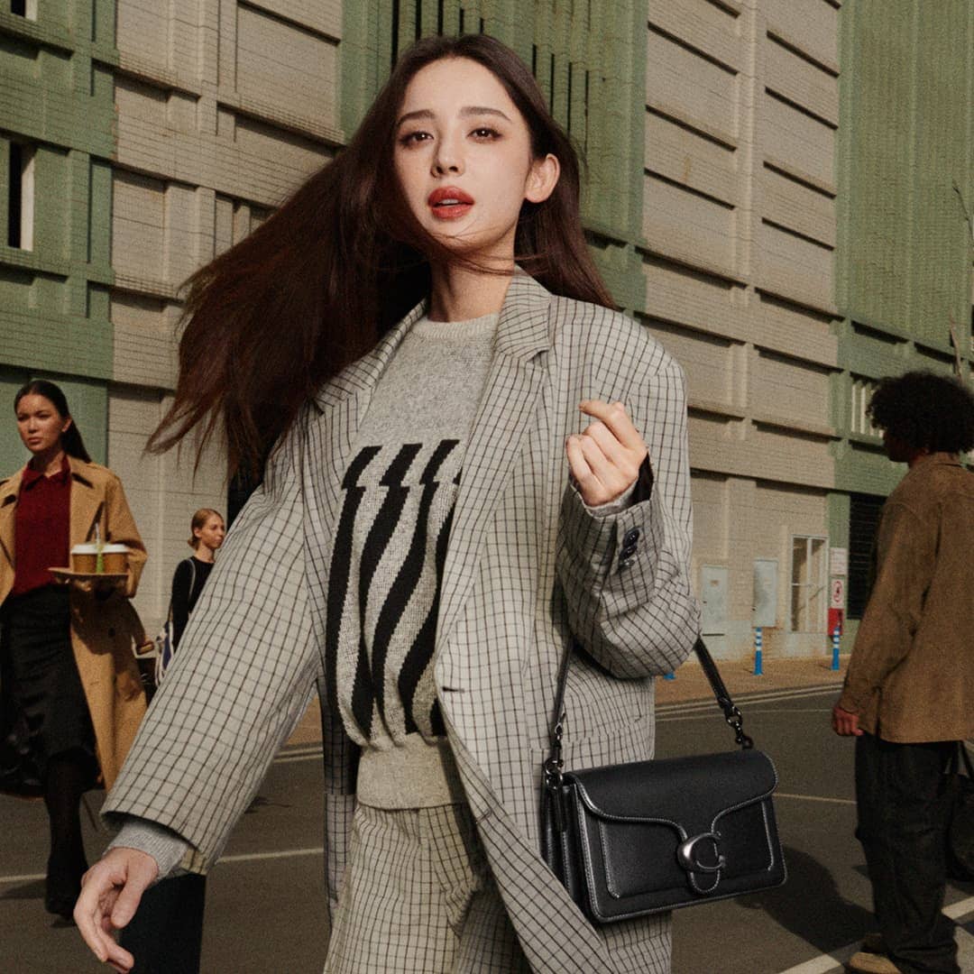 Woman in grey plaid blazer wearing a black shoulder bag walking outdoors with city buildings in background