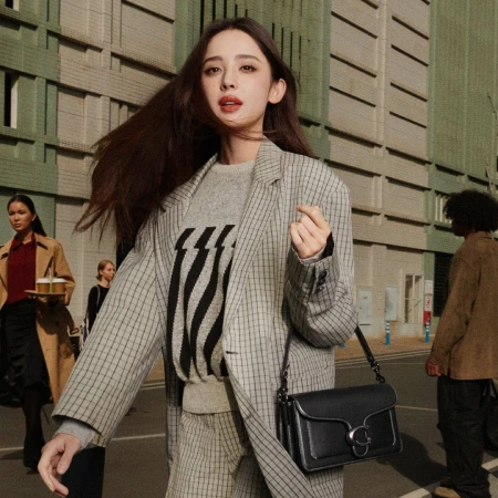 Woman in grey plaid blazer wearing a black shoulder bag walking outdoors with city buildings in background