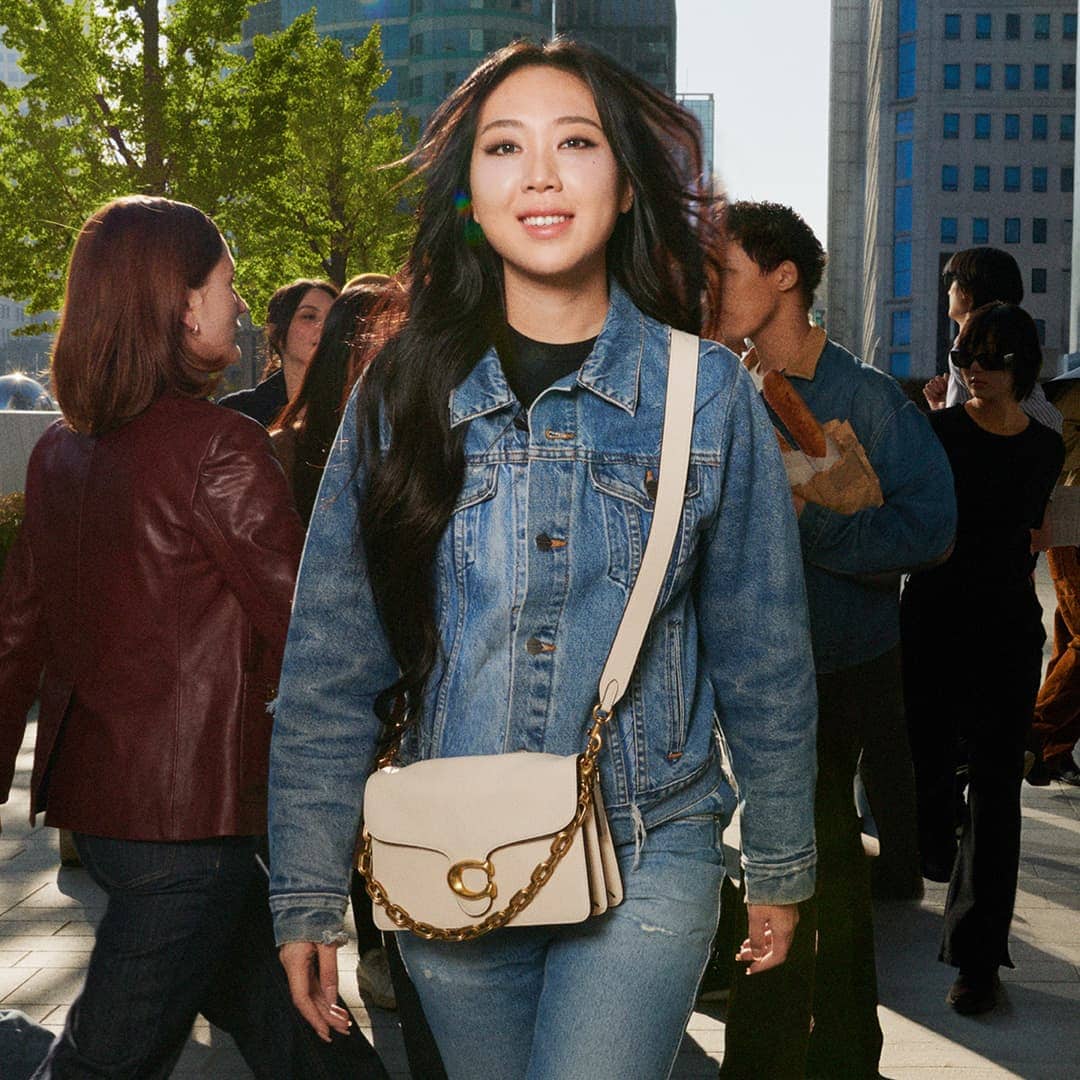 Youngji Lee wearing a denim jacket and crossbody handbag outdoors on a sunlit city street for Coach Spring 2025 campaign