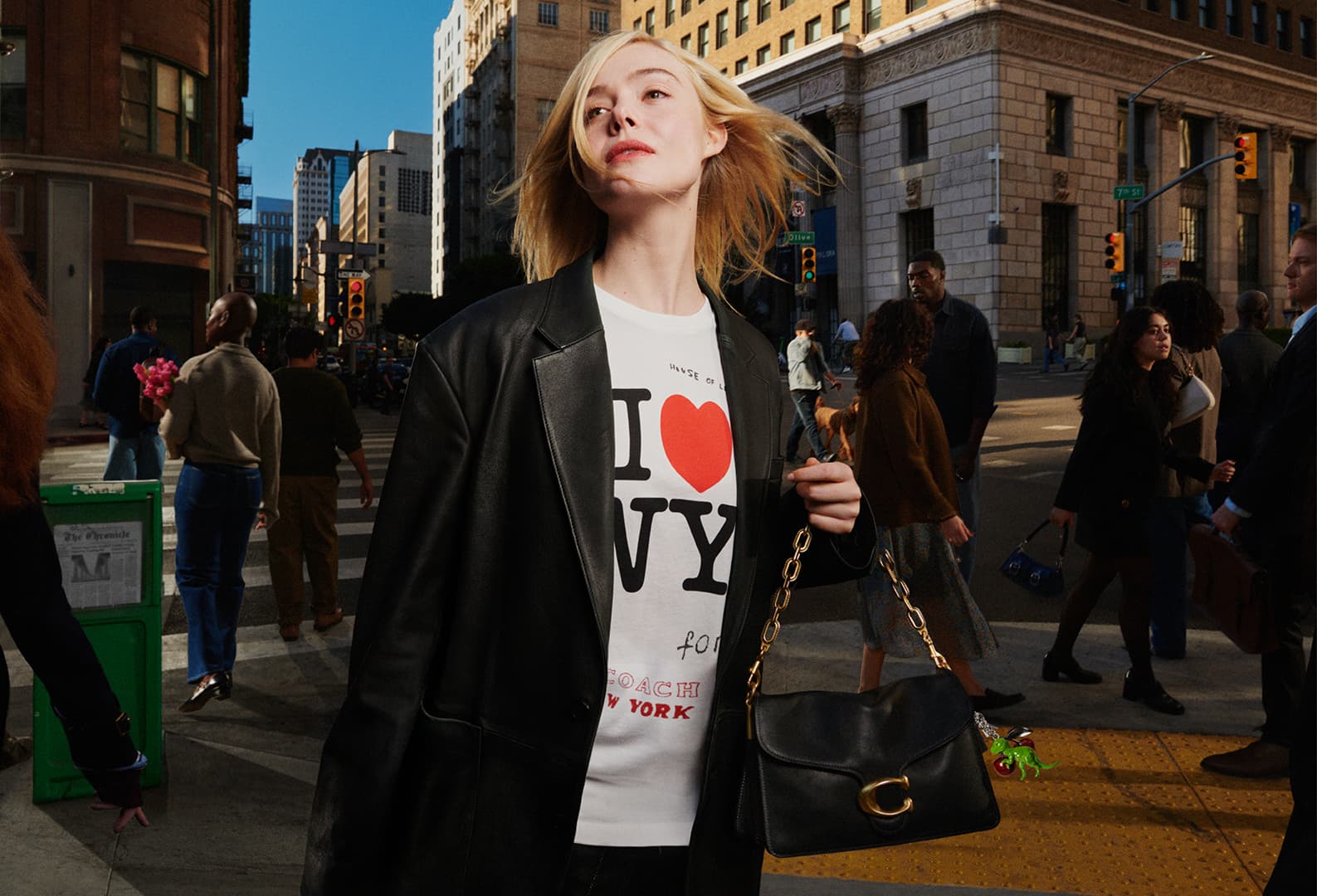 Elle Fanning in a black leather blazer and graphic t-shirt holding a black handbag on a city street.