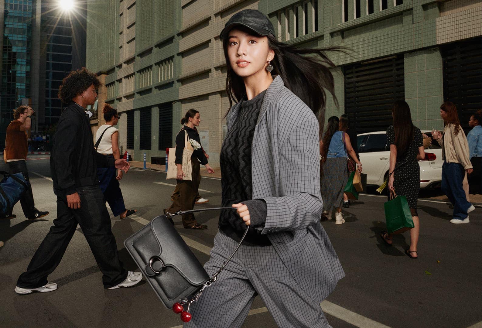 Model in grey plaid suit and cap walking down a city street holding a black leather handbag