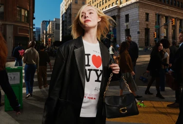 Elle Fanning in a black leather blazer and graphic t-shirt holding a black handbag on a city street.