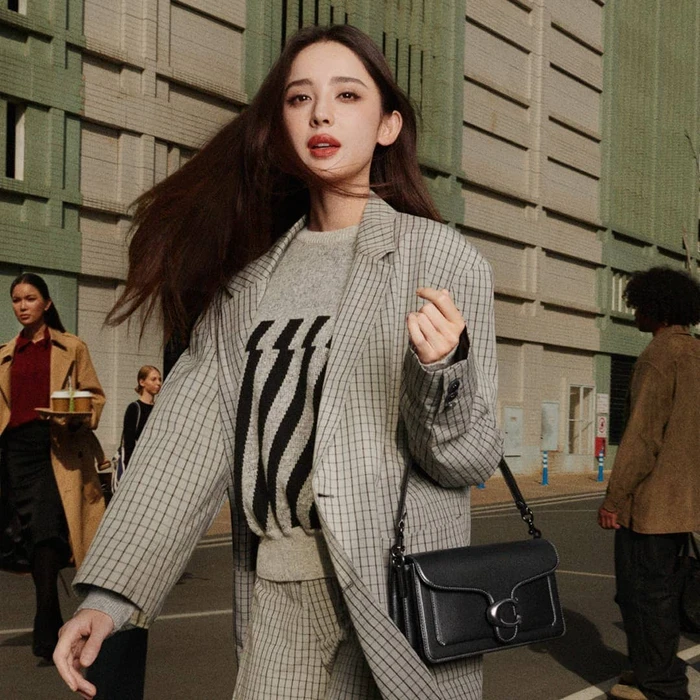 Woman in grey plaid blazer wearing a black shoulder bag walking outdoors with city buildings in background