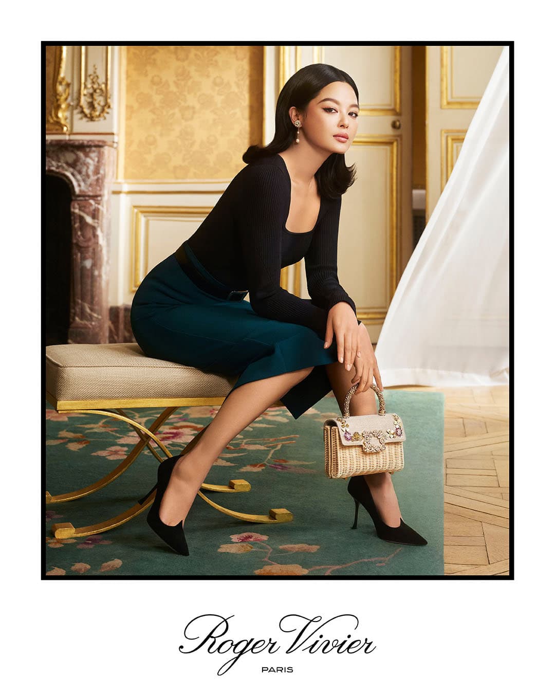 Woman in dark long-sleeve top and teal skirt seated on an upholstered bench holding a structured straw handbag in a luxury interior