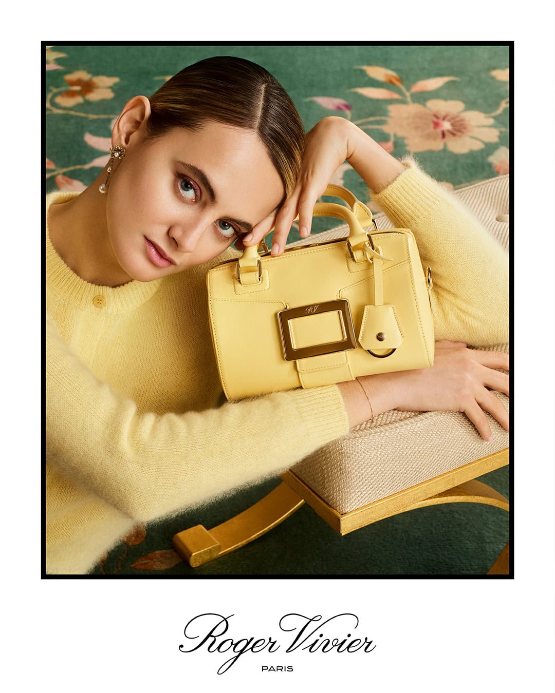 Woman in yellow sweater posing with a yellow leather handbag resting on an upholstered bench over a patterned carpet