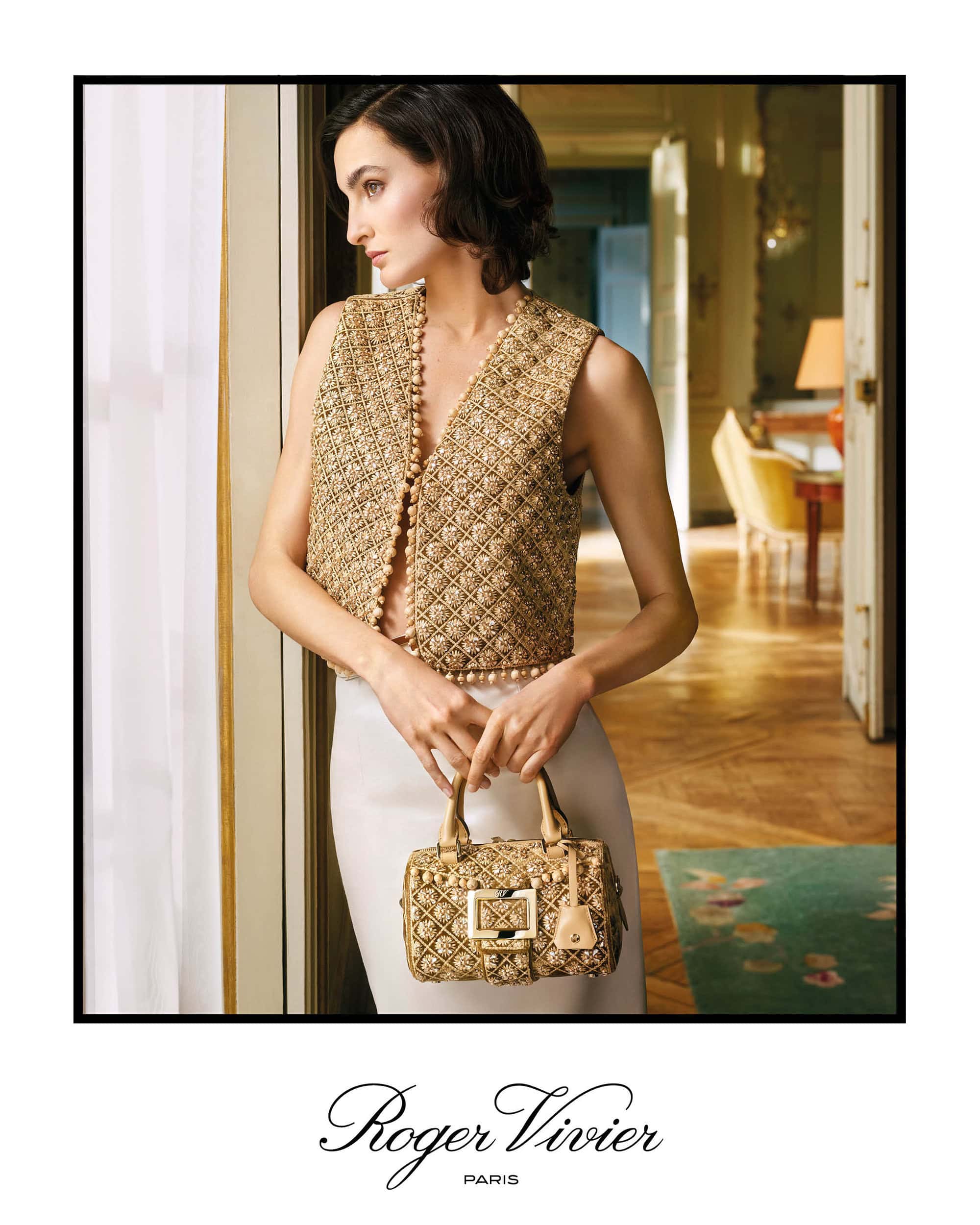 Model in a beaded gold vest and white skirt holding a matching handbag inside a sunlit room for Roger Vivier