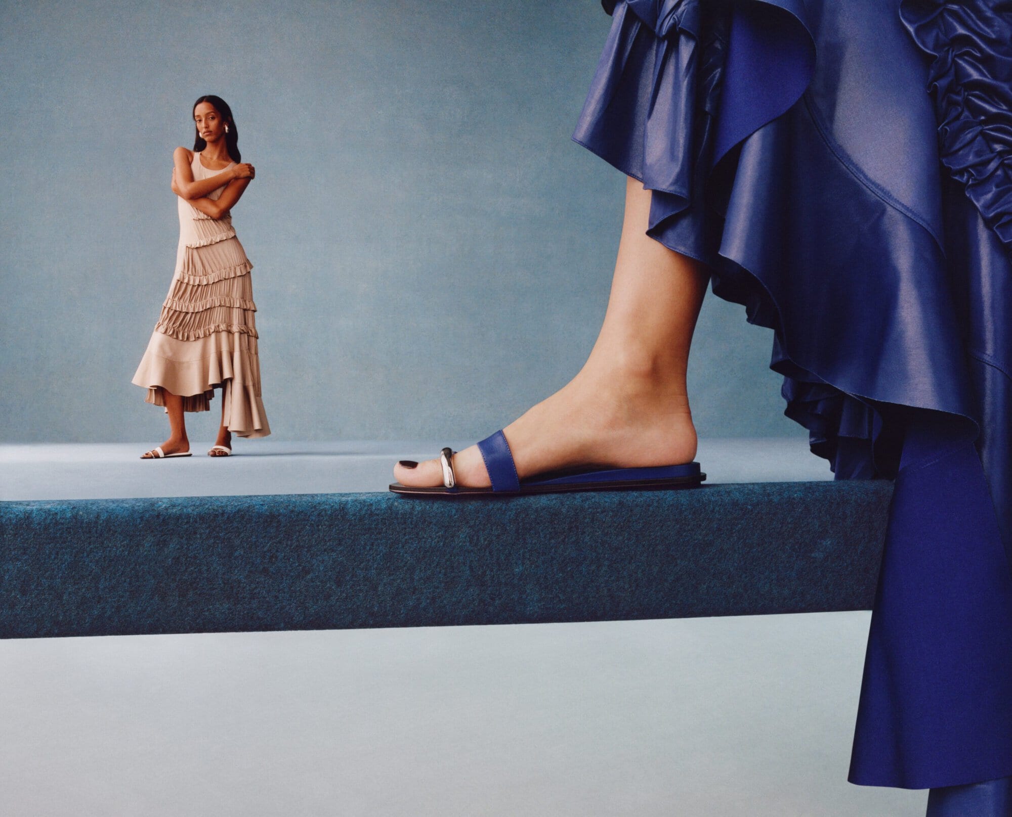 Close-up of a blue sandal on a raised platform, with a model standing in the background against a blue wall.