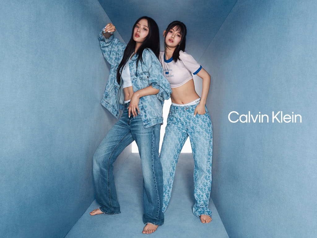 Two women posing in an angular blue corridor wearing Calvin Klein denim and white crop tops for the Spring 2025 campaign.