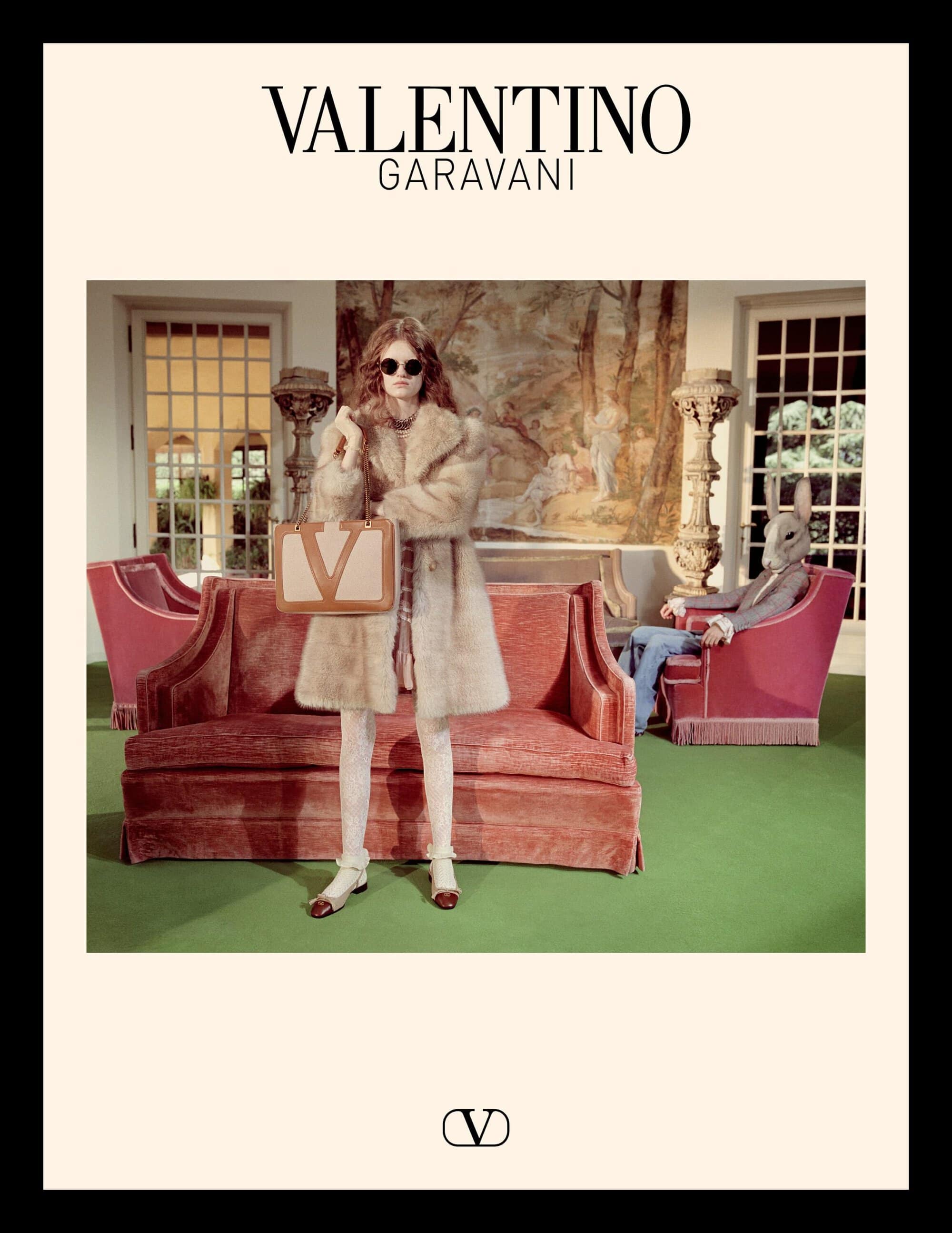 Model wearing a faux fur coat holding a tan handbag in a lavish room with a seated figure wearing a rabbit mask