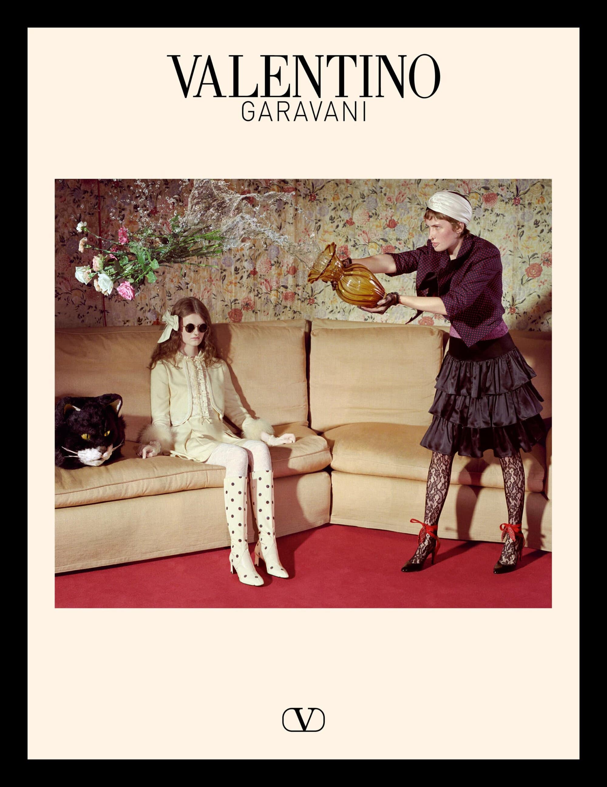 Model seated on a beige sofa wearing a light yellow dress while another person pours water from an amber vase