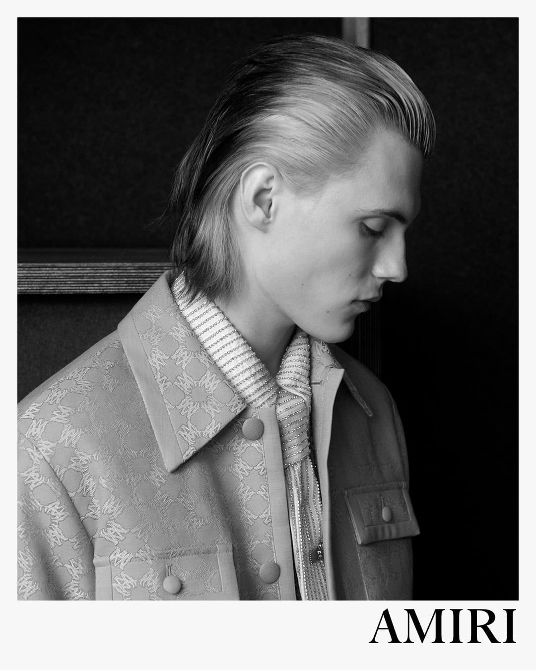 Black and white portrait of a male model in a patterned jacket and textured scarf against a dark background for Amiri