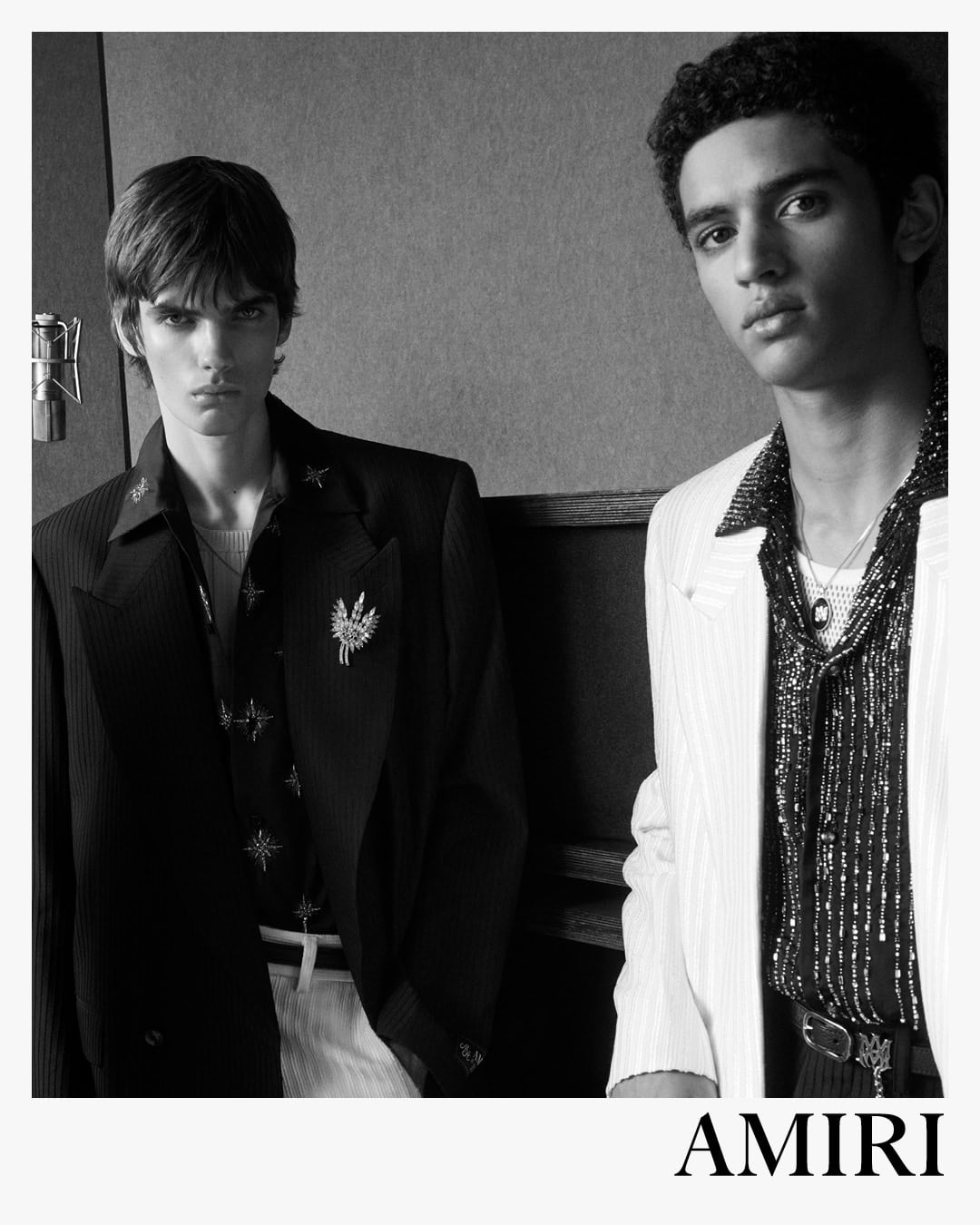 Two male models in tailored blazers and patterned shirts photographed in black and white against a textured interior background.