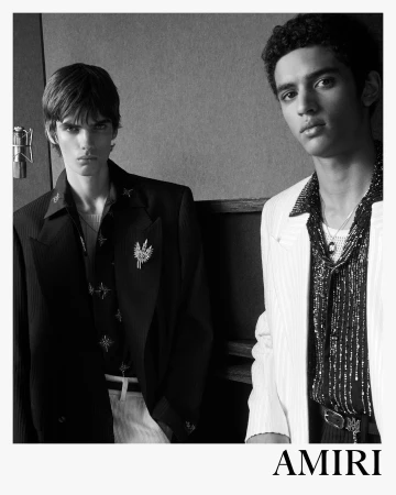 Two male models in tailored blazers and patterned shirts photographed in black and white against a textured interior background.