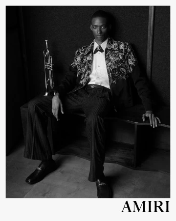 Black and white fashion campaign image featuring a model seated next to a trumpet, wearing an embroidered jacket and patterned trousers.