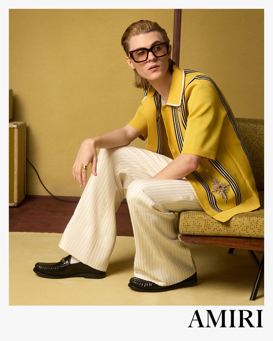 Model wearing a yellow knit polo and cream trousers seated on an armchair against a retro wall for Amiri