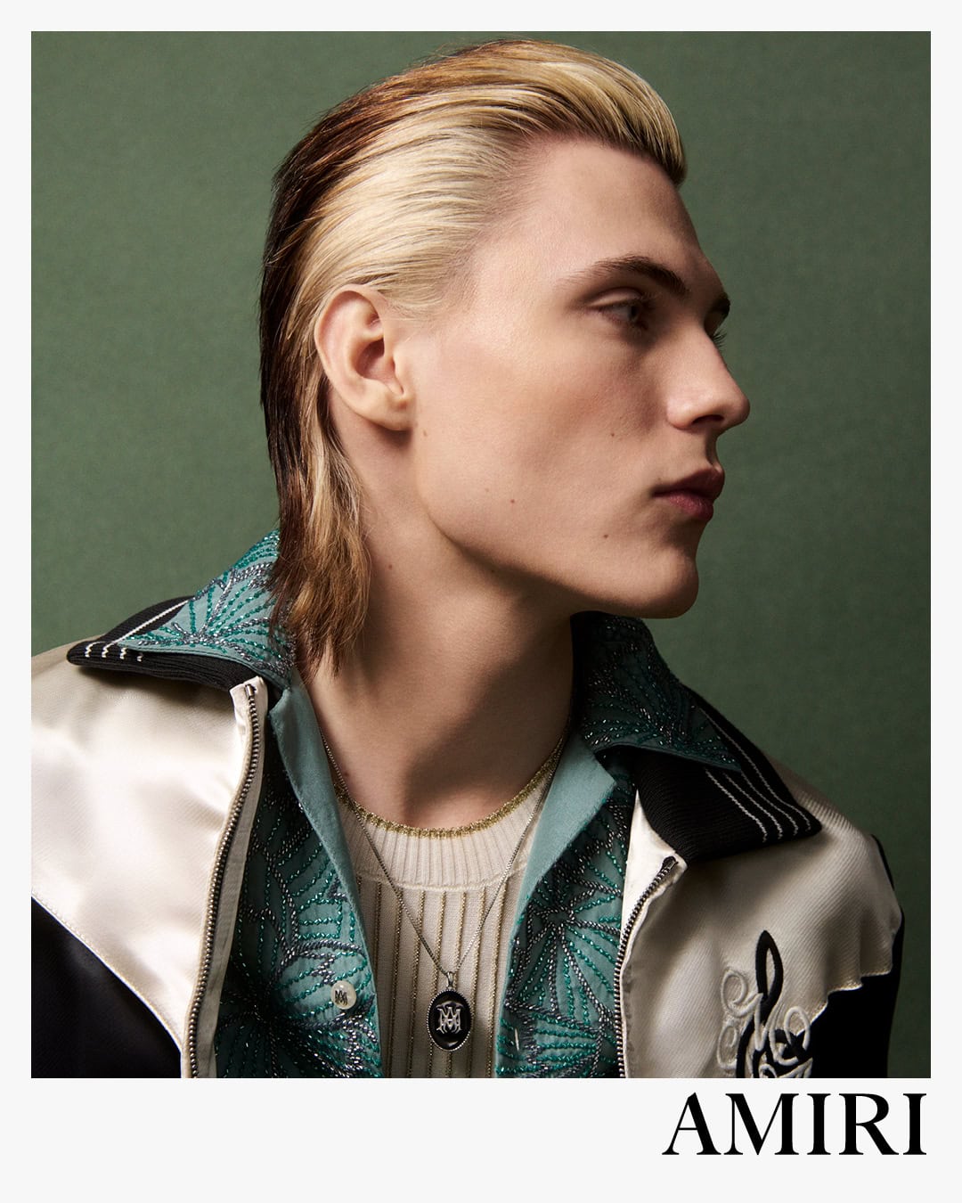 Close-up portrait of a male model with slicked-back blonde and brown hair wearing a patterned jacket and layered necklaces against a green wall
