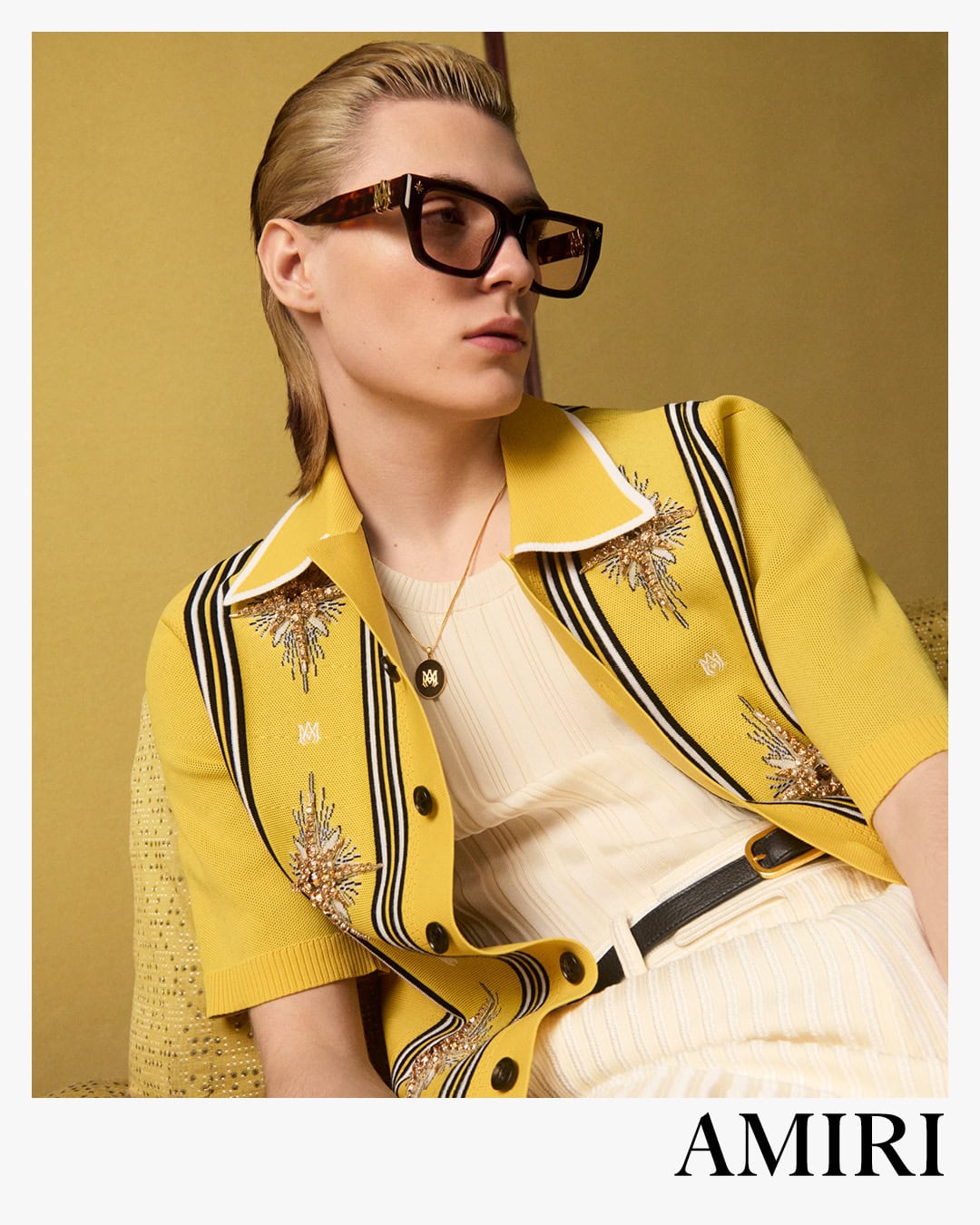 Model with slicked-back hair wearing tortoiseshell sunglasses and a yellow embroidered knit shirt against a yellow wall