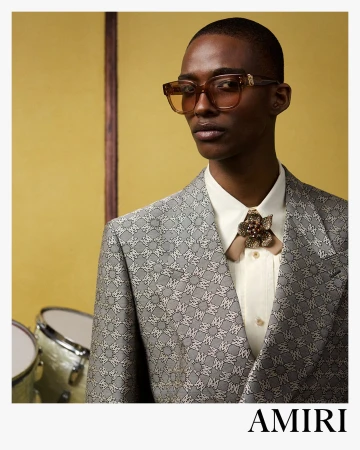 Model wearing patterned grey blazer and tinted sunglasses with an AMIRI logo on white bottom border
