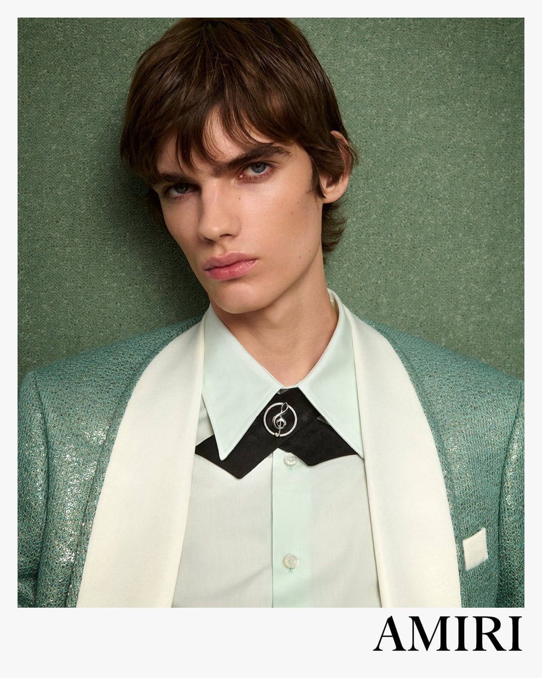 Portrait of a male model wearing a mint green jacket, white collared shirt, and black necktie with silver accessory against textured green background