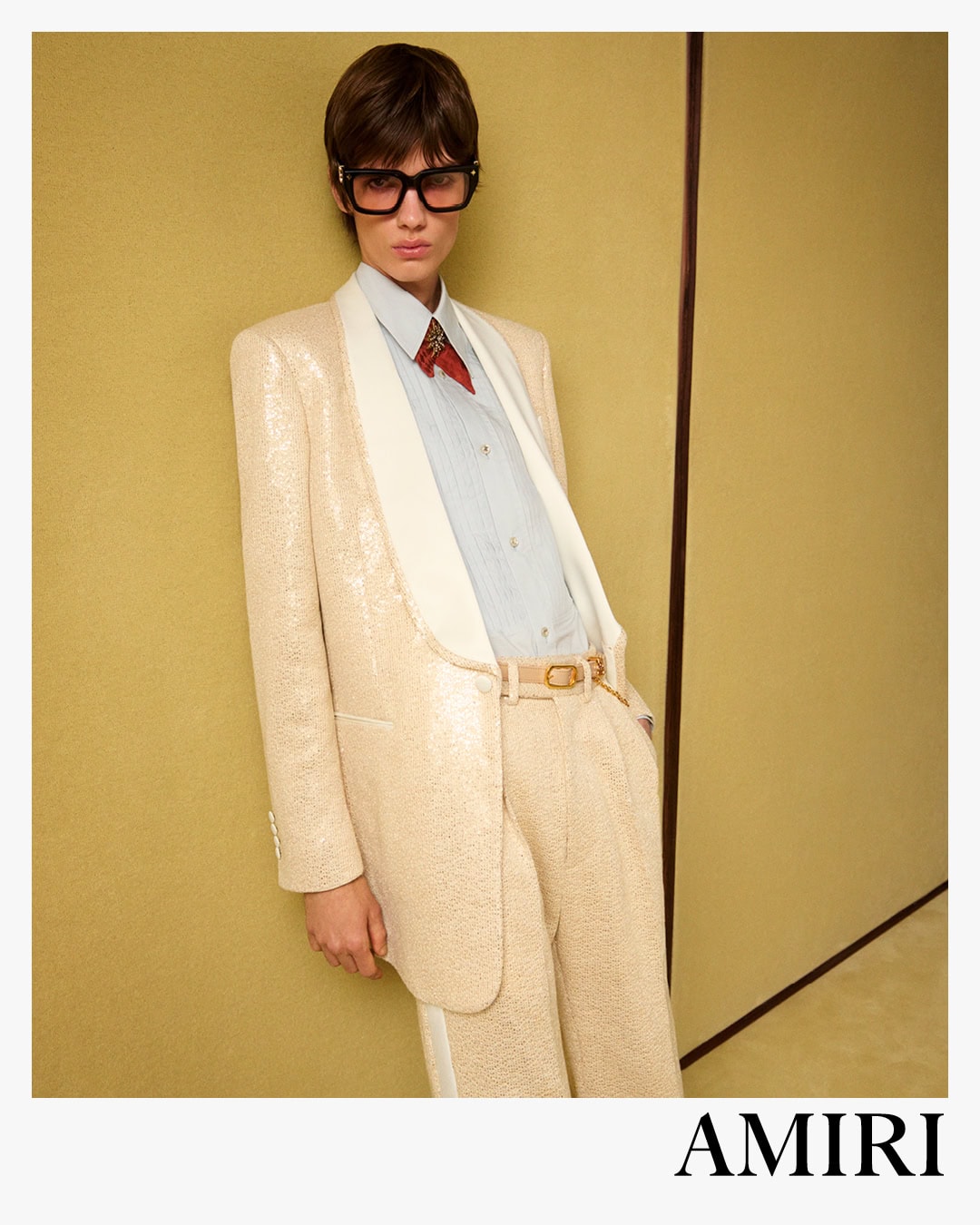 Model wearing a sequined cream suit jacket and trousers with oversized black sunglasses against a warm yellow wall