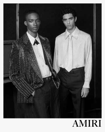 Monochrome fashion campaign portrait of two male models in tailored designer suits against a dark textured background