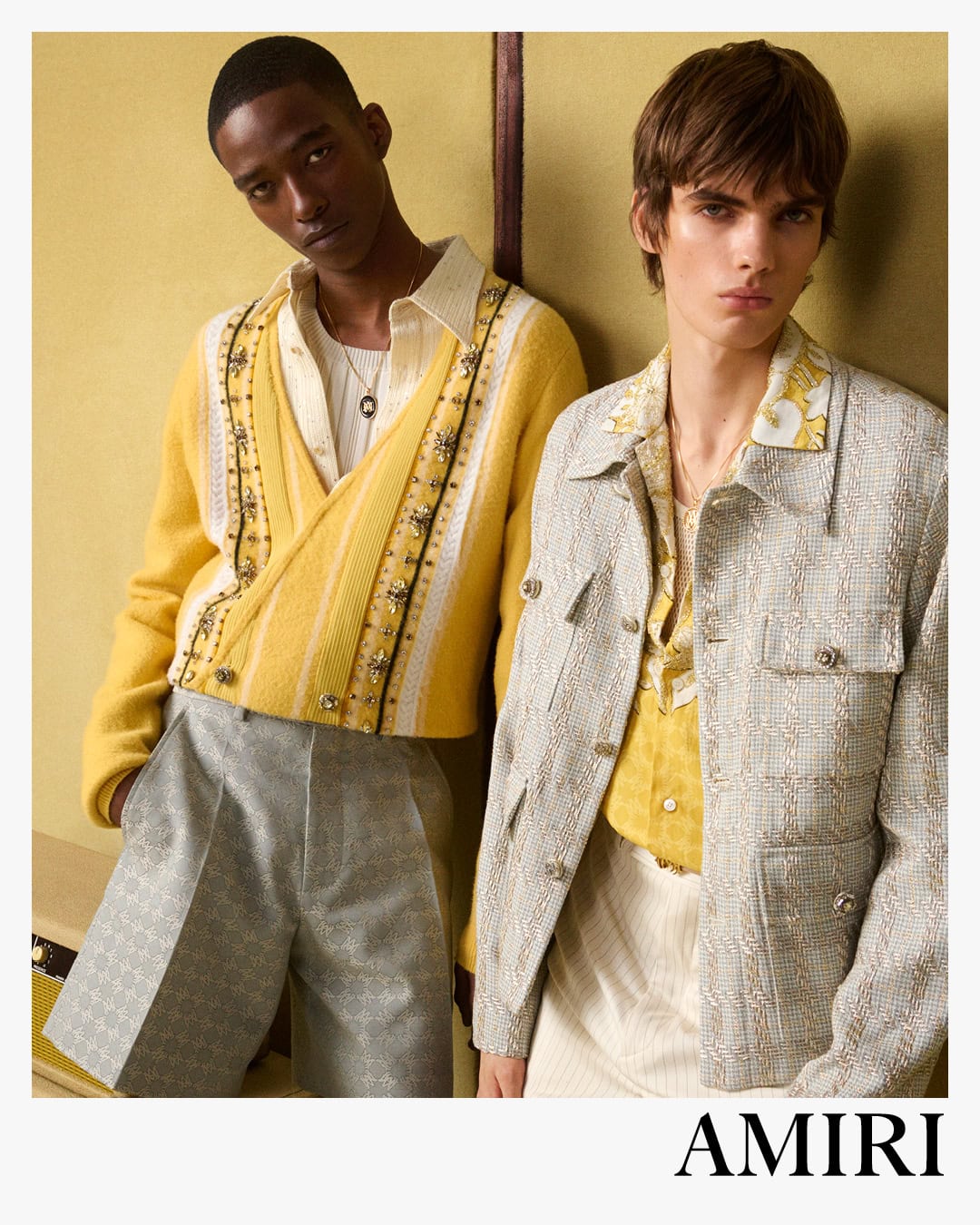 Two male models posing against a panelled wall for the Amiri Spring 2025 campaign, wearing textured knitwear and patterned jackets.